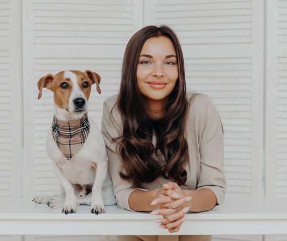 A woman with long brown hair smiles warmly, leaning on a white table next to a Jack Russell Terrier wearing a plaid bandana.