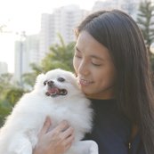 A person smiling and holding a fluffy white dog outdoors, with city buildings and trees in the background